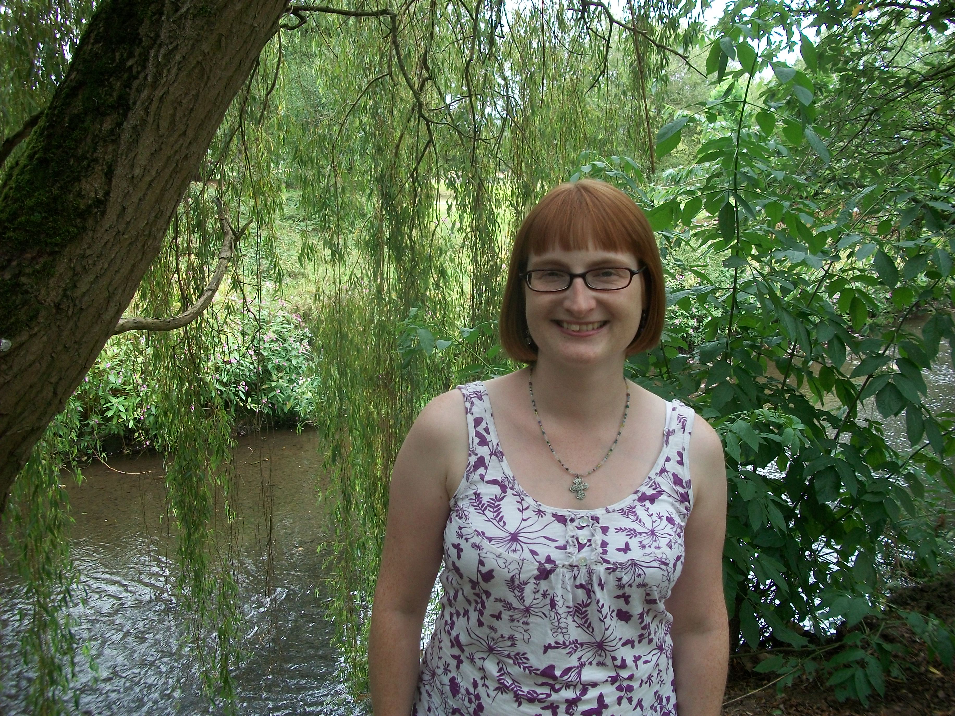 A woman with short ginger hair wearing glasses and a floral top, standing next to a river beneath a weeping willow tree. Photo of Paranormal Author Catherine Green copyright SpookyMrsGreen.com pagan lifestyle blog.