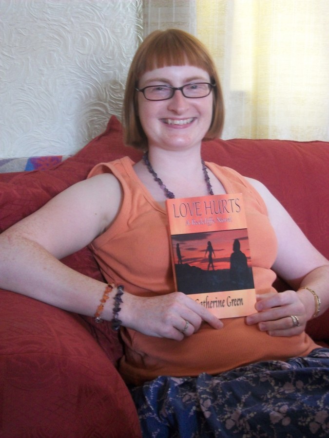 A woman with short ginger hair wearing glasses, an orange vest top and purple floral skirt, sitting on a red sofa and holding a book called Love Hurts by Catherine Green. Photo of Paranormal Author Catherine Green copyright SpookyMrsGreen.com pagan lifestyle blog.