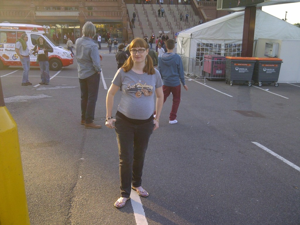 Photo of a pregnant woman with long ginger hair, wearing glasses, a Bon Jovi t-shirt, black jeans and flipflops, standing in the carpark of Aston Villa football stadium. Copyright Catherine Green of SpookyMrsGreen.com pagan lifestyle blog.