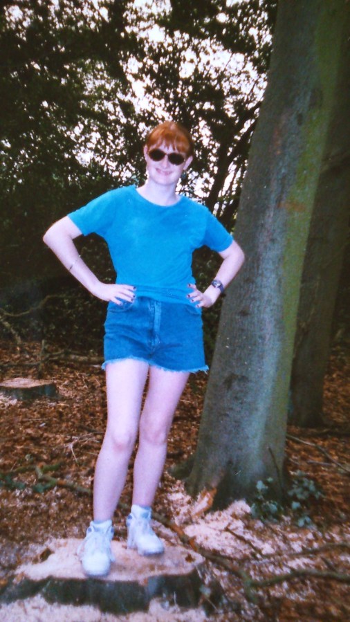 Photo of a teenage girl with ginger hair tied up in a pony tail, wearing sunglasses, a blue t-shirt, blue denim shorts, and white trainers, standing on a tree stump in a forest. Copyright Catherine Green of SpookyMrsGreen.com pagan lifestyle blog.