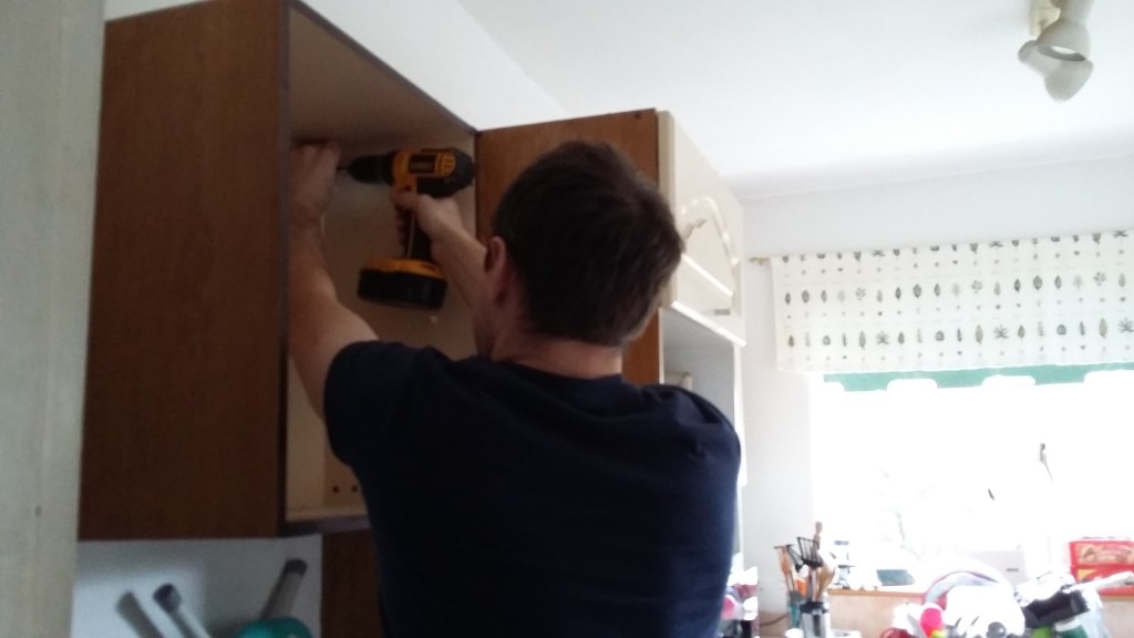 Image of a man with a drill removing wall cupboards in the small kitchen of a Victorian terraced house in UK. My Kitchen Renovation Project: Bye Bye Old Cupboards! at SpookyMrsGreen.com mindful parenting and modern pagan lifestyle blog.
