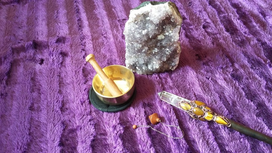 Image of a purple blanket with a large amethyst crystal geode, a small Tibetan singing bowl, a goldstone pendulum and a clear quartz and Tiger's Eye crystal wand. Crystal meditation article by Catherine Green for SpookyMrsGreen.com mindful parenting and modern pagan lifestyle blog.