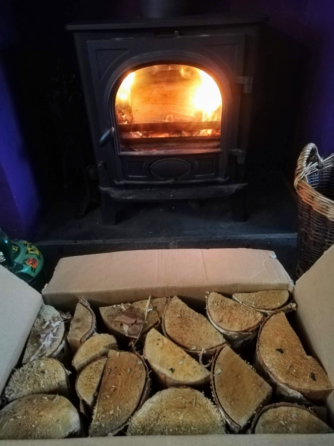 Image of a log fire with a cosy fire burning and a box of logs in front of the fire. #ProductReview White Horse Energy logs and kindling reviewed by Catherine Green at SpookyMrsGreen.com mindful parenting and modern pagan lifestyle blog.