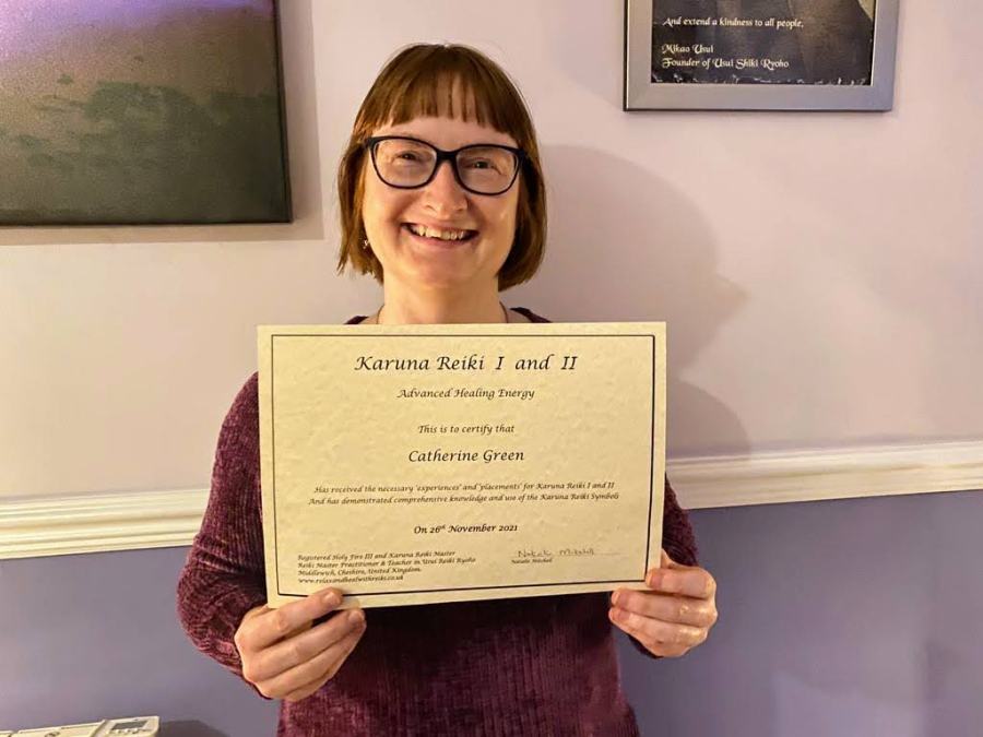 Photo of Catherine Green from Goldstone Reiki holding a certificate for Karuna Reiki level 1 and 2. Catherine has short ginger hair and wears glasses and a purple jumper. She is smiling.