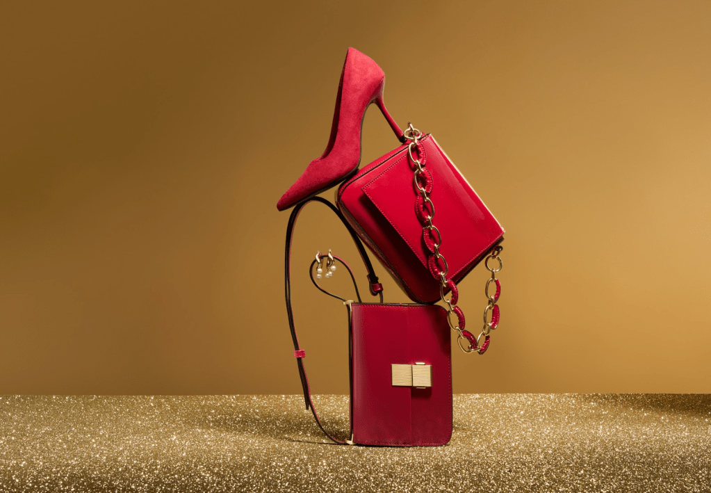 Gold glitter background with image of a red stiletto shoe balancing on red handbags with gold chain handles. Festive Sale at M&S with Black Friday sales on Clinique, 20% off fashion, gifts and furniture plus lots more huge deals affiliated with SpookyMrsGreen.com mindful parenting and modern pagan lifestyle blog.