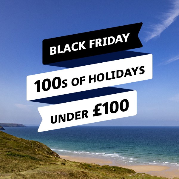 Tranquil image of a sandy beach with the sea gently lapping against the sand with blue sky above and green grass on the cliff. Text reads, "Black Friday Hundreds of holidays under one hundred pounds." Haven Holidays affiliated with SpookyMrsGreen.com mindful parenting and modern pagan lifestyle blog.