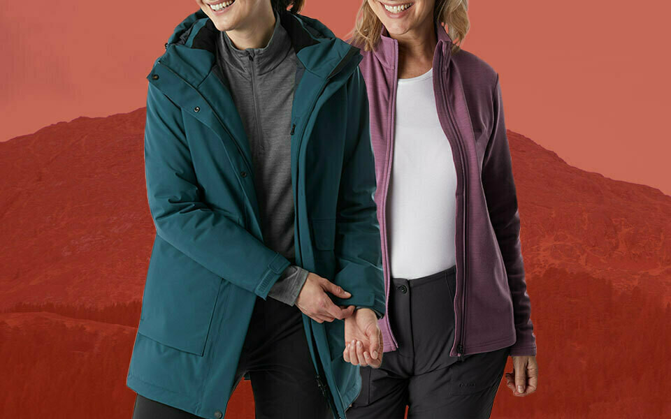 Red background with distant mountains. Image of a woman wearing a blue walking coat with grey fleece jumper and jeans and a woman wearing a purple fleece jacket with white top and black walking trousers. Rohan outdoor clothing affiliated with SpookyMrsGreen.com mindful parenting and modern pagan lifestyle blog.