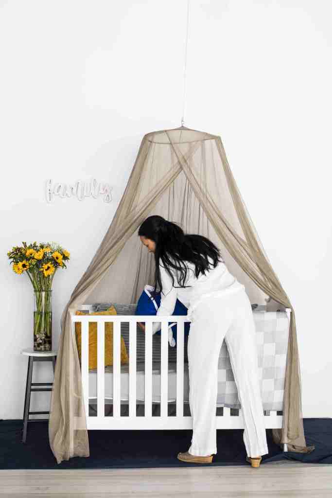 Image of a woman checking her baby in a cot that stands underneath a Shield Your Body SYB Serenity Canopy. Shield Your Body affiliated with SpookyMrsGreen.com mindful parenting and modern pagan lifestyle blog.