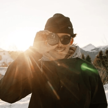 Image of a man dressed in outdoor clothing and standing in the sun with mountains behind him, holding Natural Sunscreen by Palm & Pine affiliated with SpookyMrsGreen mindful parenting and modern pagan lifestyle blog.