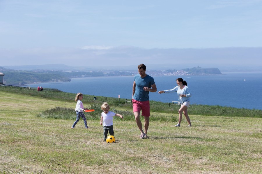 Image of a mum and dad playing football with their children in a field overlooking the sea. School Summer Holidays under 300GBP at Haven Holidays affiliated with SpookyMrsGreen.com mindful parenting and modern pagan lifestyle blog.