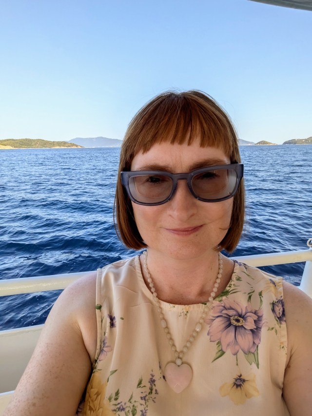 Colourful photo of a woman with red hair ginger hair wearing sunglasses Ziena glasses moisture chamber glasses with the mediterranean sea in the background. Text reads “Mummy Monday: Living with Rare Eye Disease and Chronic Pain.” UK pagan family pagan mom at SpookyMrsGreen.com spooky family lifestyle blog.